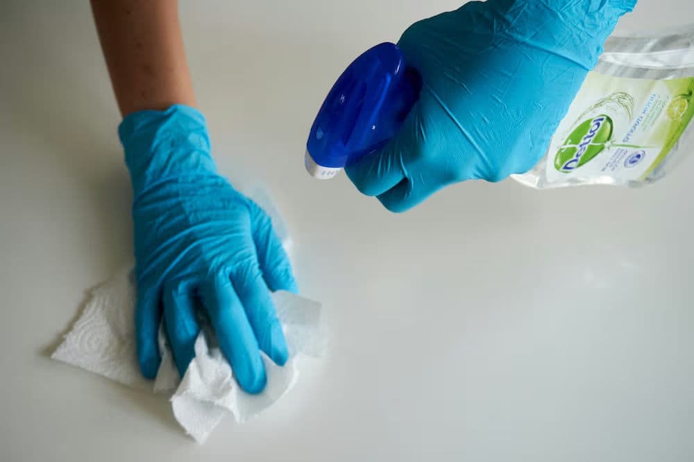 Person in blue gloves spraying cleaner on a surface and wiping with paper towel.